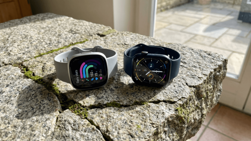 Fitbit Sense 2 vs Apple Watch Series 10 placed side by side on a stone table