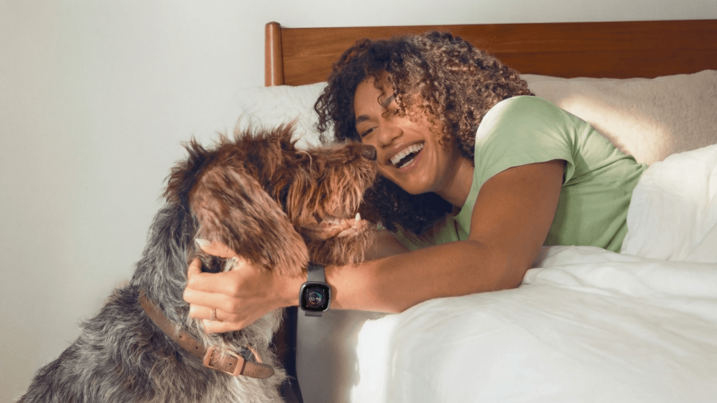 Woman wearing Fitbit Sense 2 while in bed with a dog standing close by
